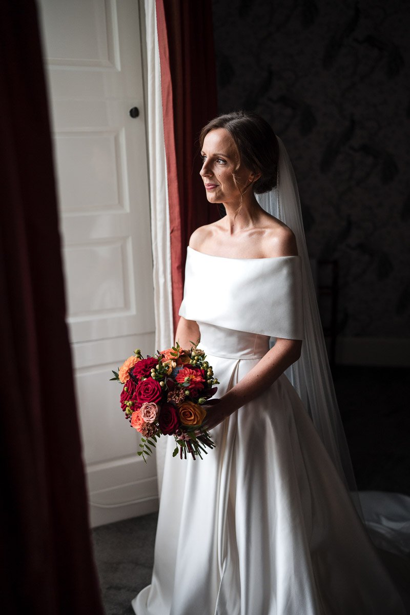A bride in a white off-shoulder wedding gown holding a colorful bouquet, standing beside a window with curtains, looking out at Boturich Loch Lomond