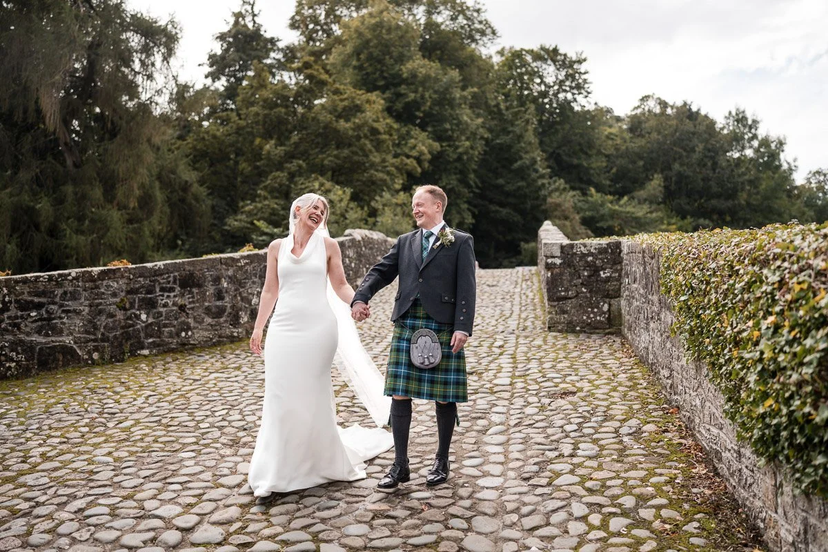 A bride and groom walking hand in hand on a cobblestone path, smiling and laughing, outdoors with trees and a stone wall in the background at Brig o' Doon