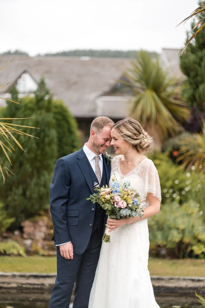 A bride and groom standing close together outdoors, smiling and touching foreheads, with the bride holding a bouquet of flowers, surrounded by lush greenery and trees at Auchrannie, Arran