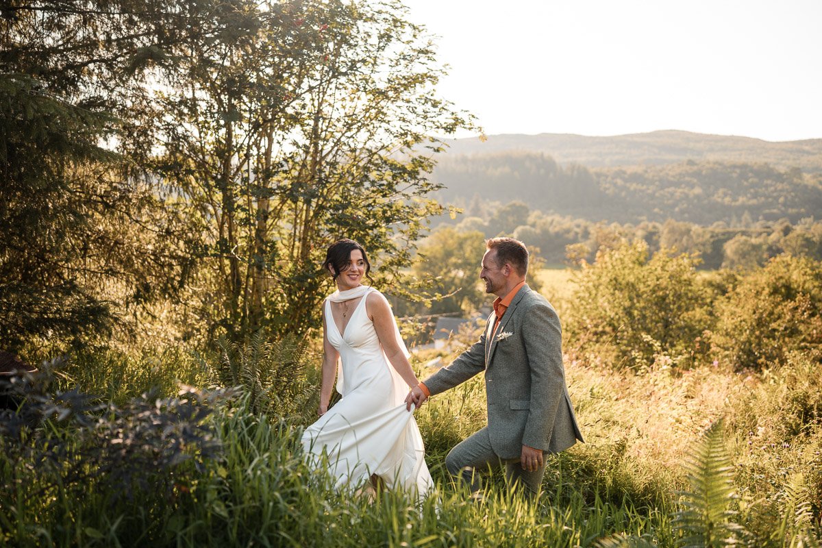 A bride and groom holding hands in a lush, green outdoor setting with trees and mountains in the background, illuminated by warm sunlight at Kilmartin Castle, Scotland