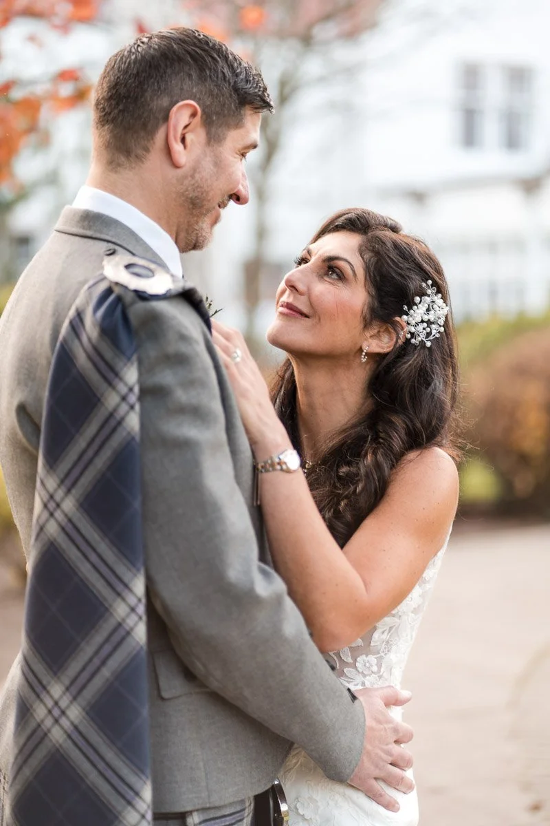 A bride and groom sharing a romantic moment outdoors, with the bride smiling up at the groom and the groom looking down at her, set against a background of trees and a white building at Lochgreen House Hotel, Troon