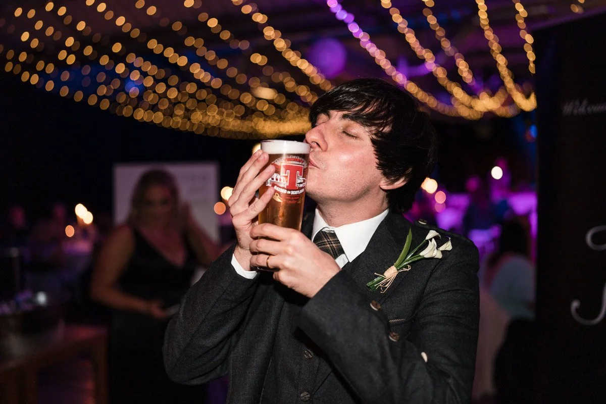 Groom in a suit and tie with a boutonniere kisses a Tennent's Lager at a wedding with string lights and a blurred background at Drygate Brewery