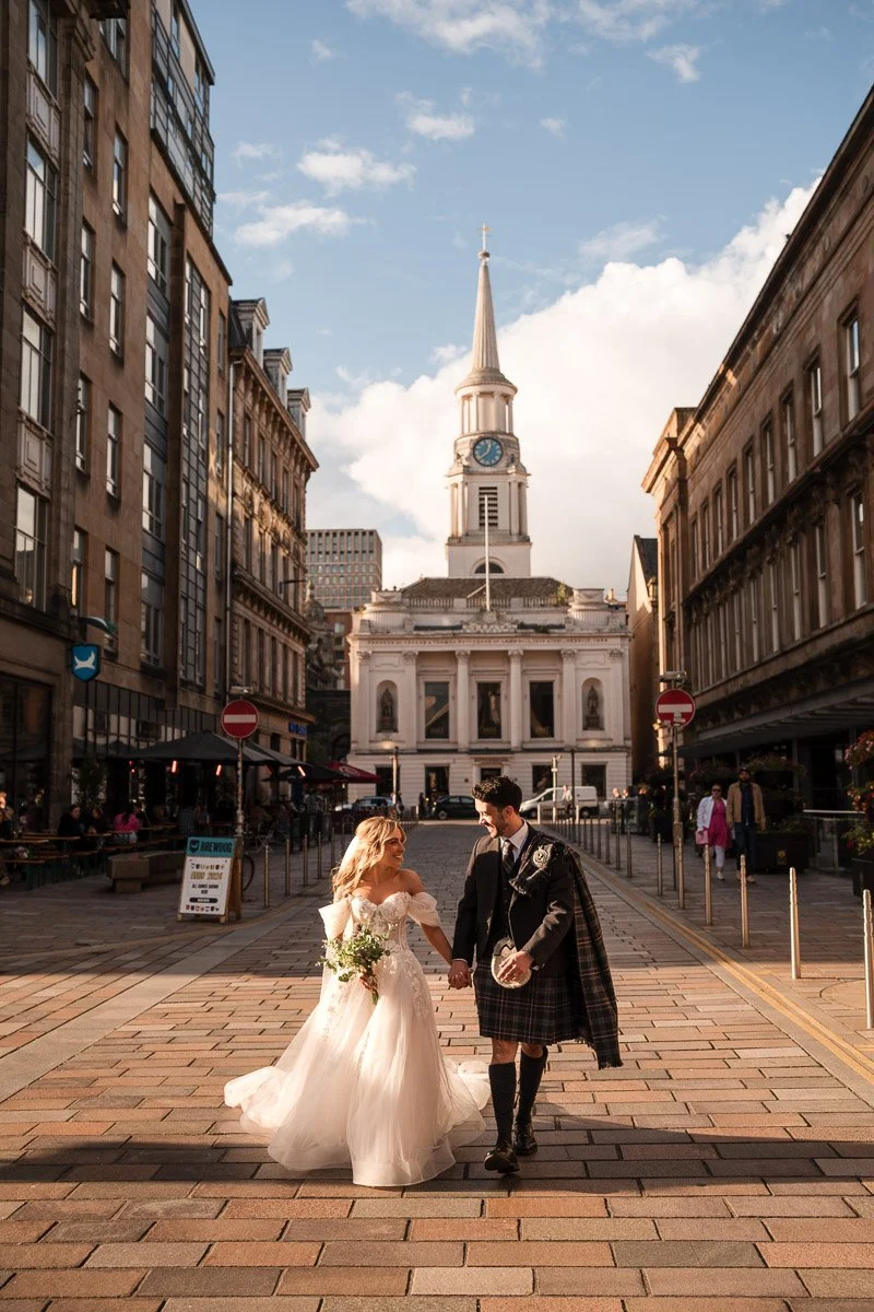 A bride and groom walking hand in hand in a city street with historic buildings and a church with a clock tower in the background at a Glasgow City elopement