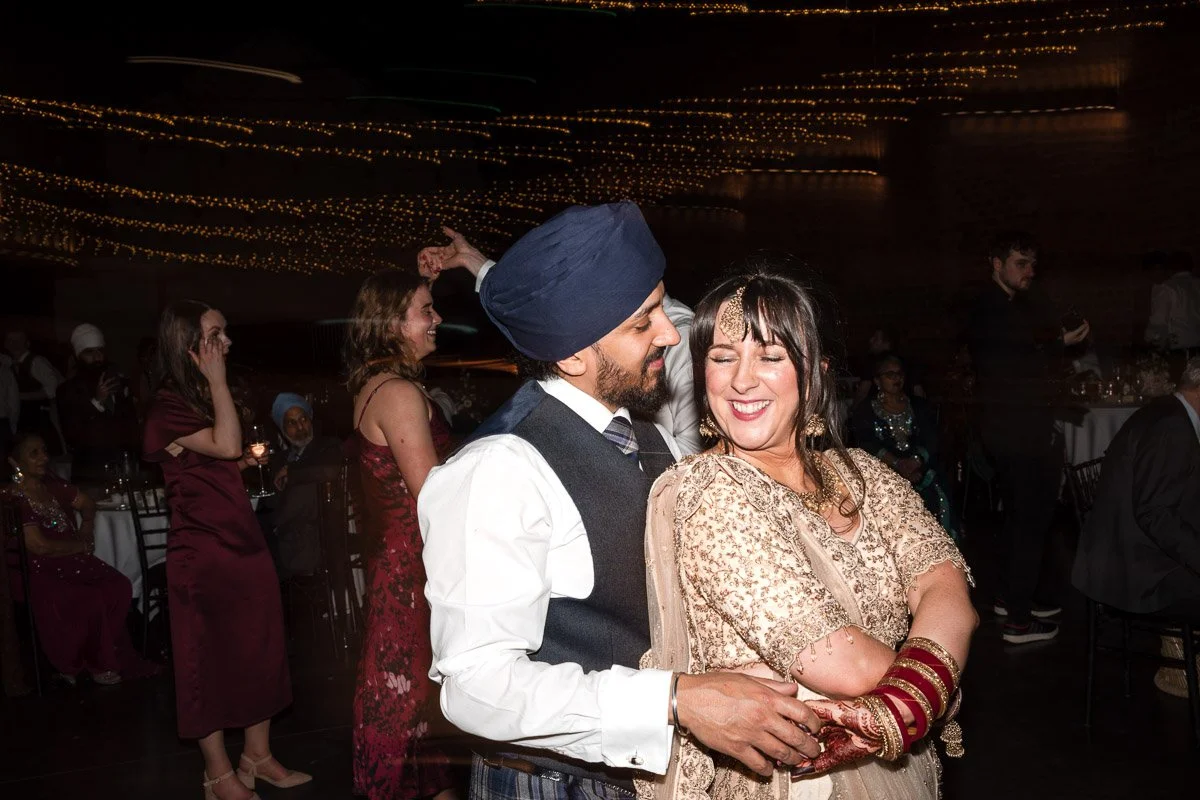 A bride and groom dancing closely at a wedding in Engine Works, with other guests watching and smiling in the background. The man wears a turban, vest, and white shirt, and the woman is dressed in a traditional embellished dress with bangles.