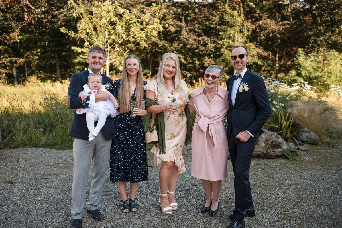 A group of six people standing outdoors in a garden, smiling at the camera, dressed in formal and semi-formal attire, during a wedding in Kilmartin Castle