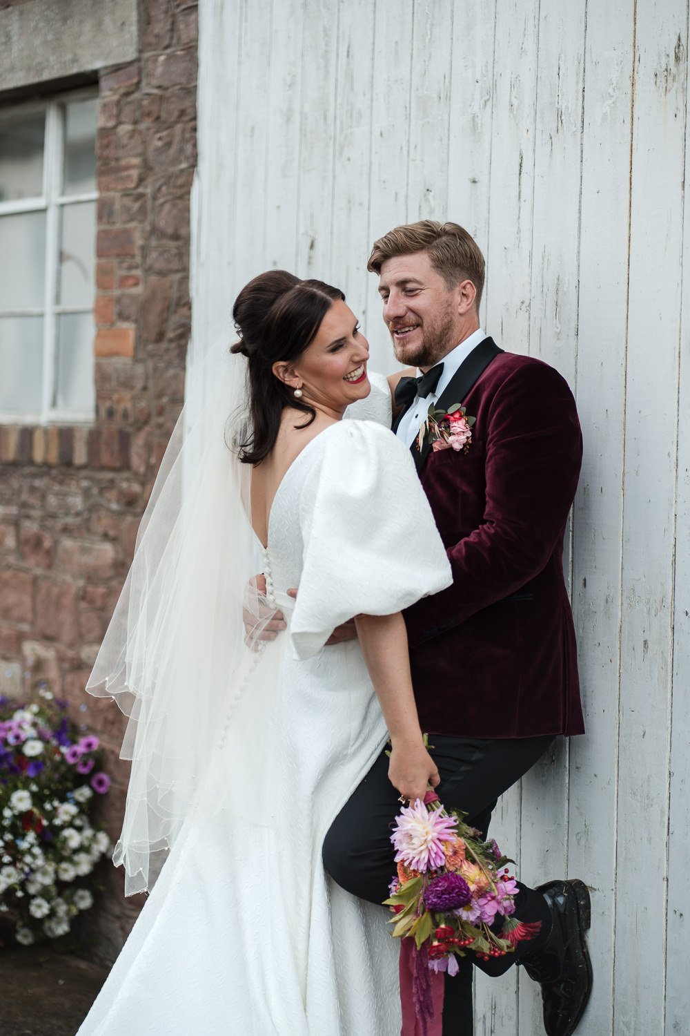 Bride and groom smiling and embracing outdoors next to a white wooden wall and brick building, with flowers nearby.