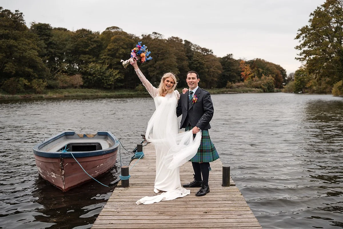 A bride and groom celebrate on a wooden dock by a loch, with the bride holding a colourful bouquet and wearing a white dress, and the groom in a suit with a kilt, both smiling at their wedding at Broxmouth Courtyard