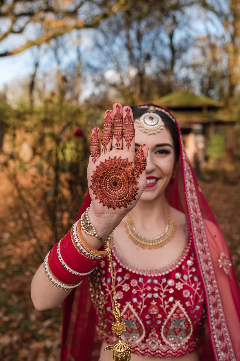 A woman dressed in traditional Indian bridal attire showing her decorated palm with henna. She is smiling, wearing jewelry, a red sari, and is outdoors with trees in the background before her Hindu wedding in Glasgow