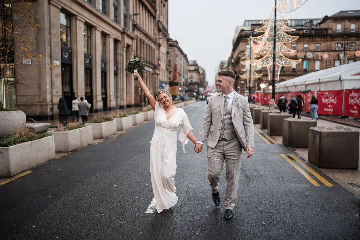 A newlywed couple walking hand in hand on a city street during a rainy evening. The bride is joyfully raising her bouquet, smiling, wearing a white wedding dress. The groom is dressed in a gray plaid suit, looking at her with a smile. Decorative holi