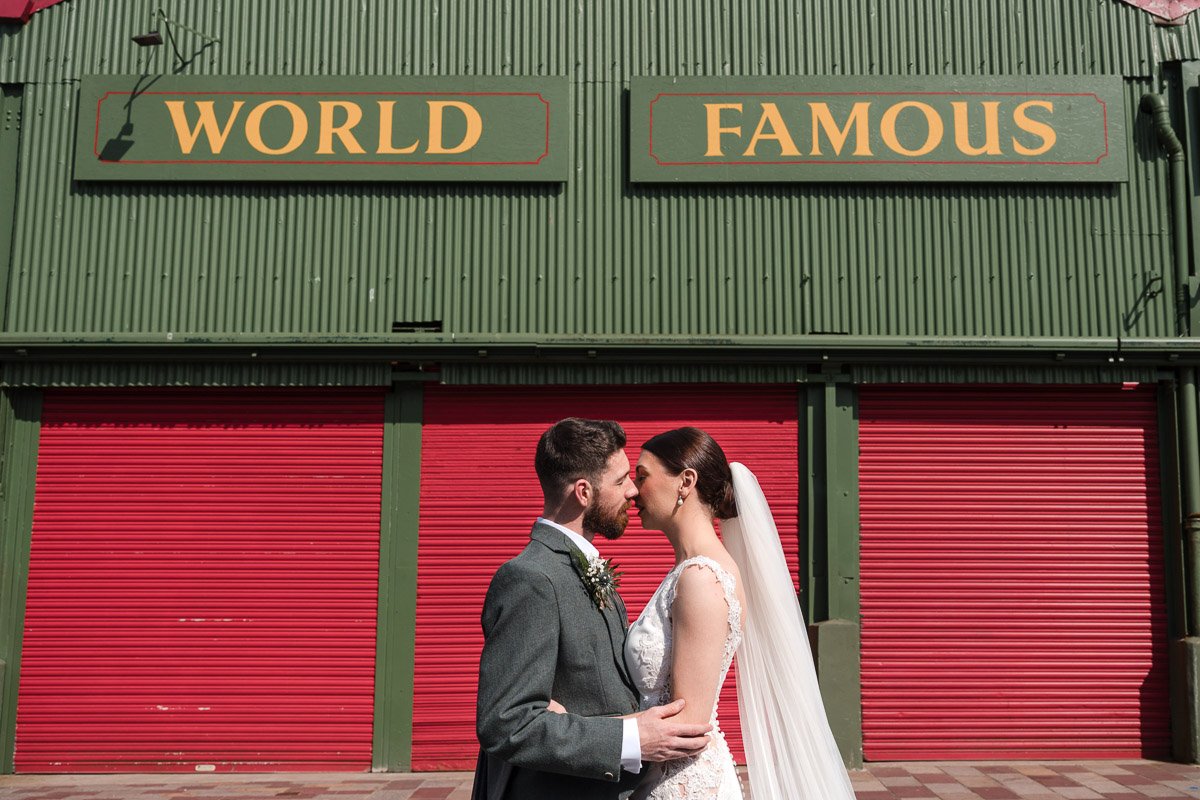 A bride and groom stand close together in front of a green building with red shutters, the sign above reads 'World Famous' at Barras Market Glasgow