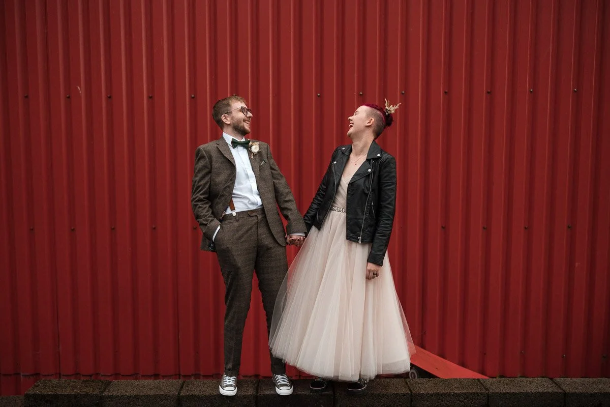 A couple holding hands, standing in front of a red metal wall, dressed in wedding attire. The man wears a brown suit with a bow tie, and the woman wears a beige tulle dress with a black leather jacket.