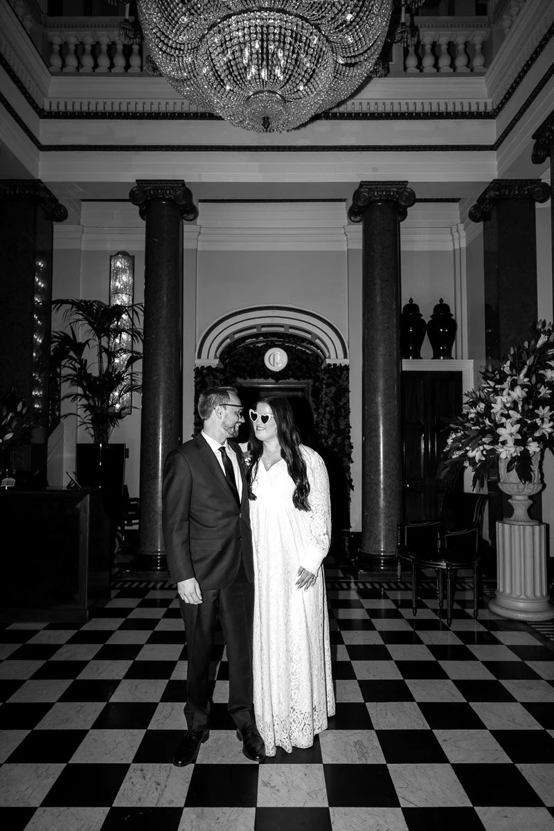 A couple dressed in formal attire standing together in an elegant, grand hall with a chandelier, tall columns, and decorative plants, smiling and looking at each other in an Edinburgh city elopement