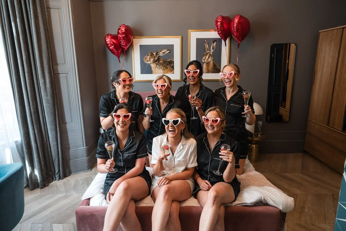 A group of seven women in pajamas celebrating with champagne and heart-shaped sunglasses during bridal prep, with heart-shaped balloons in a cozy room with animal artwork on the wall.