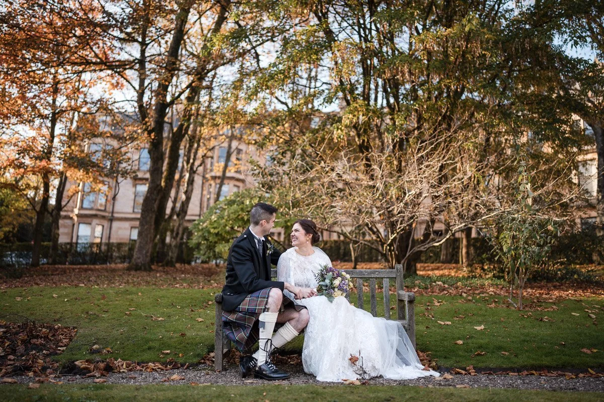 A bride and groom sitting on a park bench in a wooded area during autumn, holding hands, with trees showing fall foliage in the background after their Glasgow City elopement