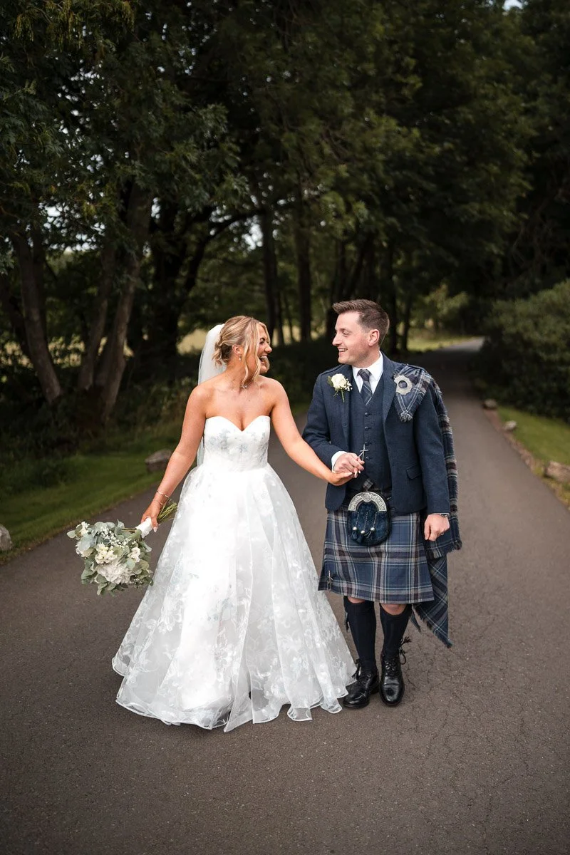 Bride in a white wedding dress holding a bouquet, smiling at groom dressed in traditional Scottish attire with kilt, walking on a paved path surrounded by trees at Cornhill Castle