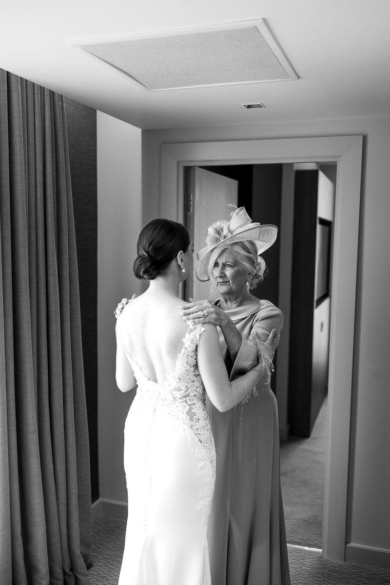 An mother of the bride in a fancy dress and large hat embraces a bride in a lace wedding gown in a hotel room before a Glasgow city elopement