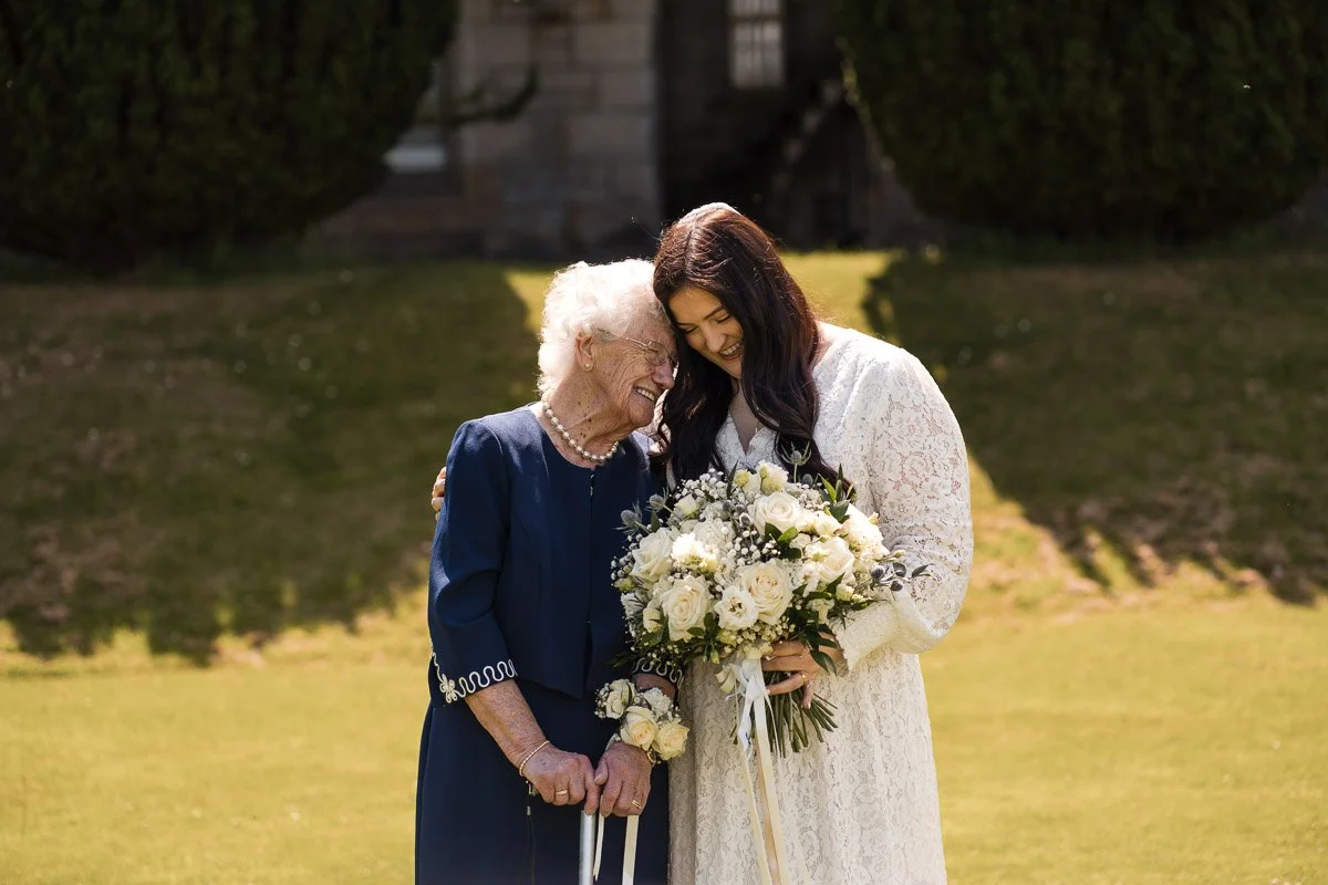 A young bride in a white lace dress holding a bouquet of white flowers in Edinburgh, standing close and laughing with an elderly woman in a navy dress and pearl necklace, outside on a sunny day.
