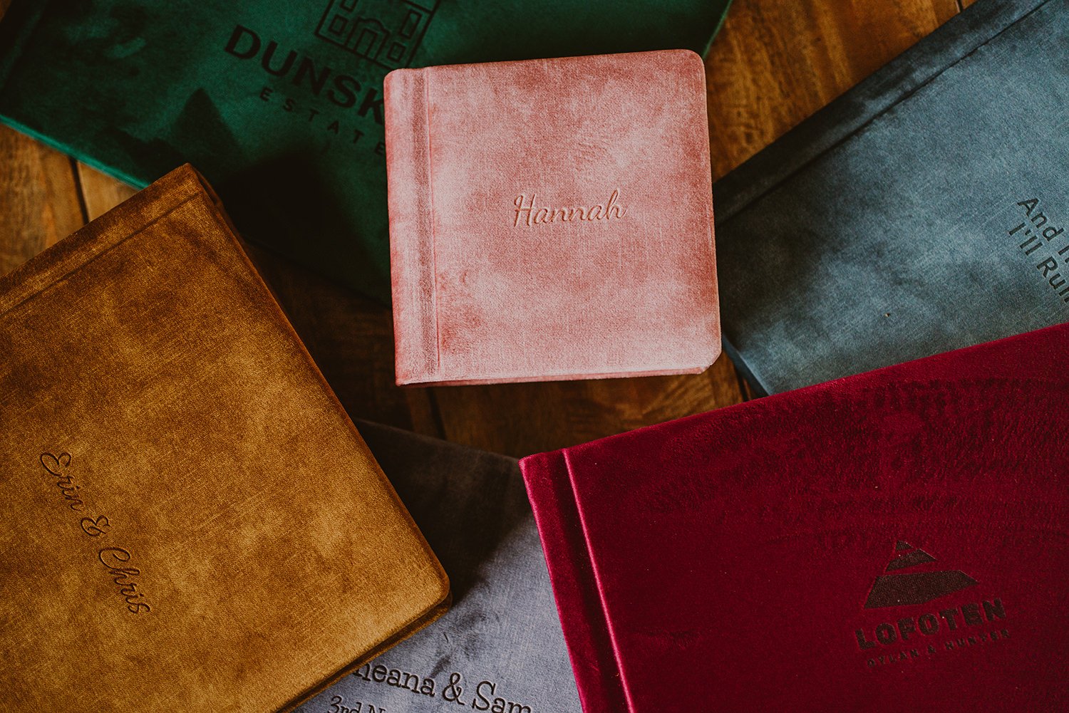 A collection of five books with different colored covers arranged on a wooden surface. The books have personalized engravings, with one titled 'Hannah' in pink, another titled 'Eli & Eli Biss' in tan, and a red book with the logo and text 'LOFOTEN DEAR & RUMER'. The other books are gray and green, with one showing the partially visible text 'And...