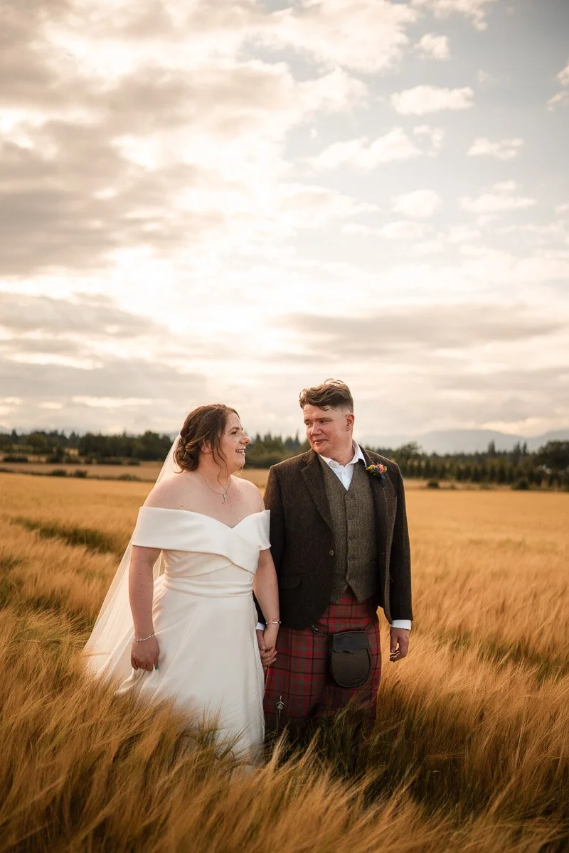 A bride and groom holding hands and walking through a field of tall grass on a cloudy day, smiling at each other.