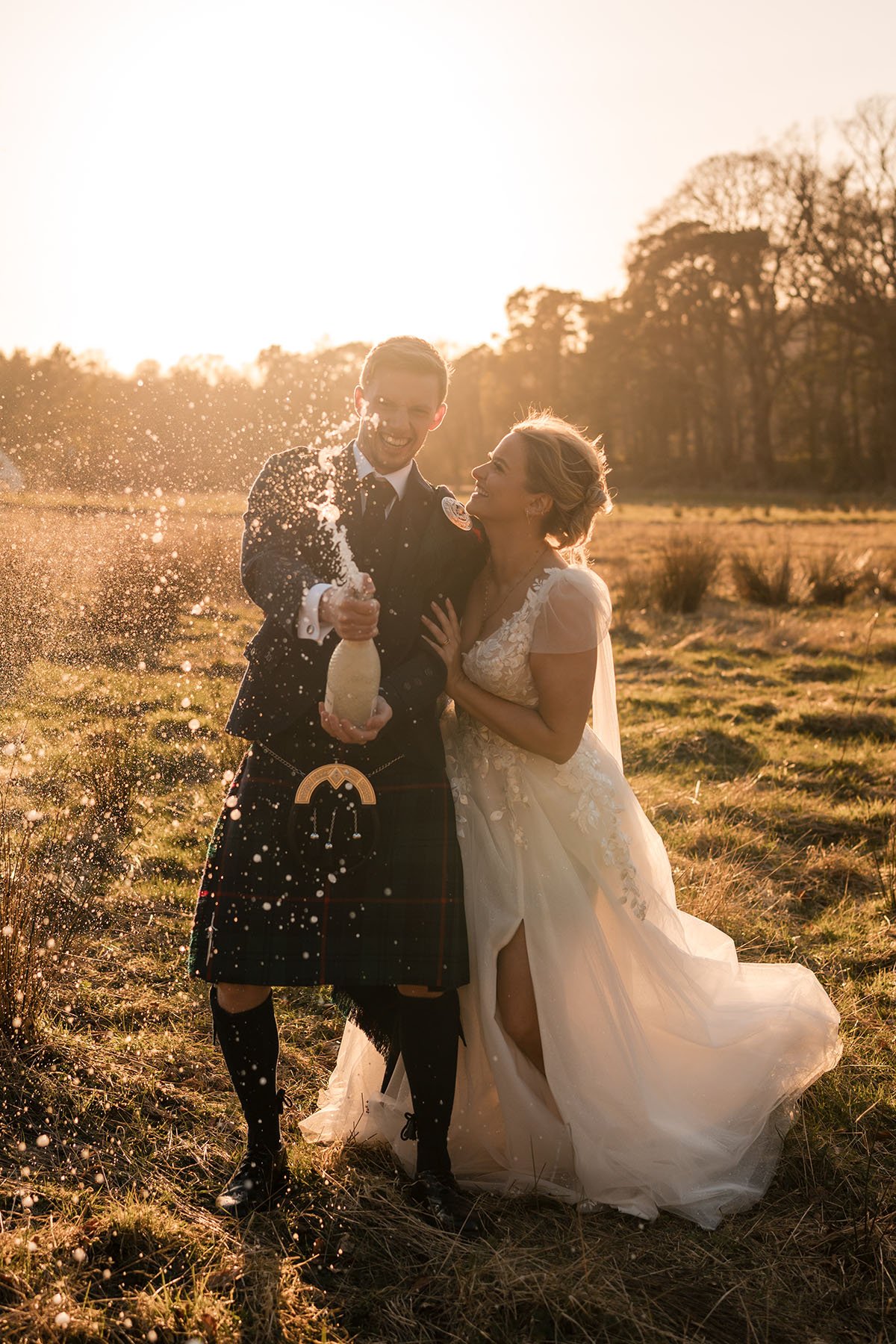 A newlywed couple celebrating outdoors at sunset, dressed in wedding attire. The groom, dressed in a kilt, is spraying champagne, and the bride, in a white wedding dress, is smiling and looking at him, at Enterkine House Hotel