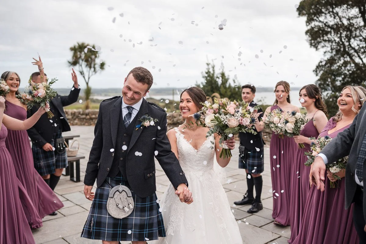 A bride and groom celebrating their wedding with friends and family outdoors, surrounded by confetti and holding hands, with women in purple dresses holding bouquets and men in kilts at GG's Yard
