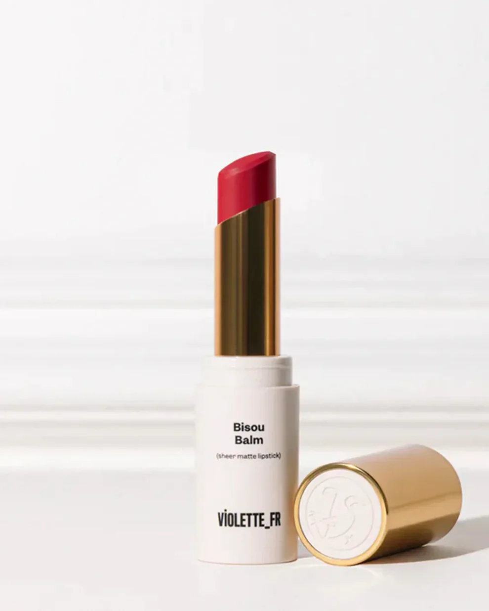 A lipstick in a white and gold tube with a red sheer matte lipstick. The tube is labeled 'Bisou Balm' and 'VIOLETTE_FR' with a gold cap lying beside it.