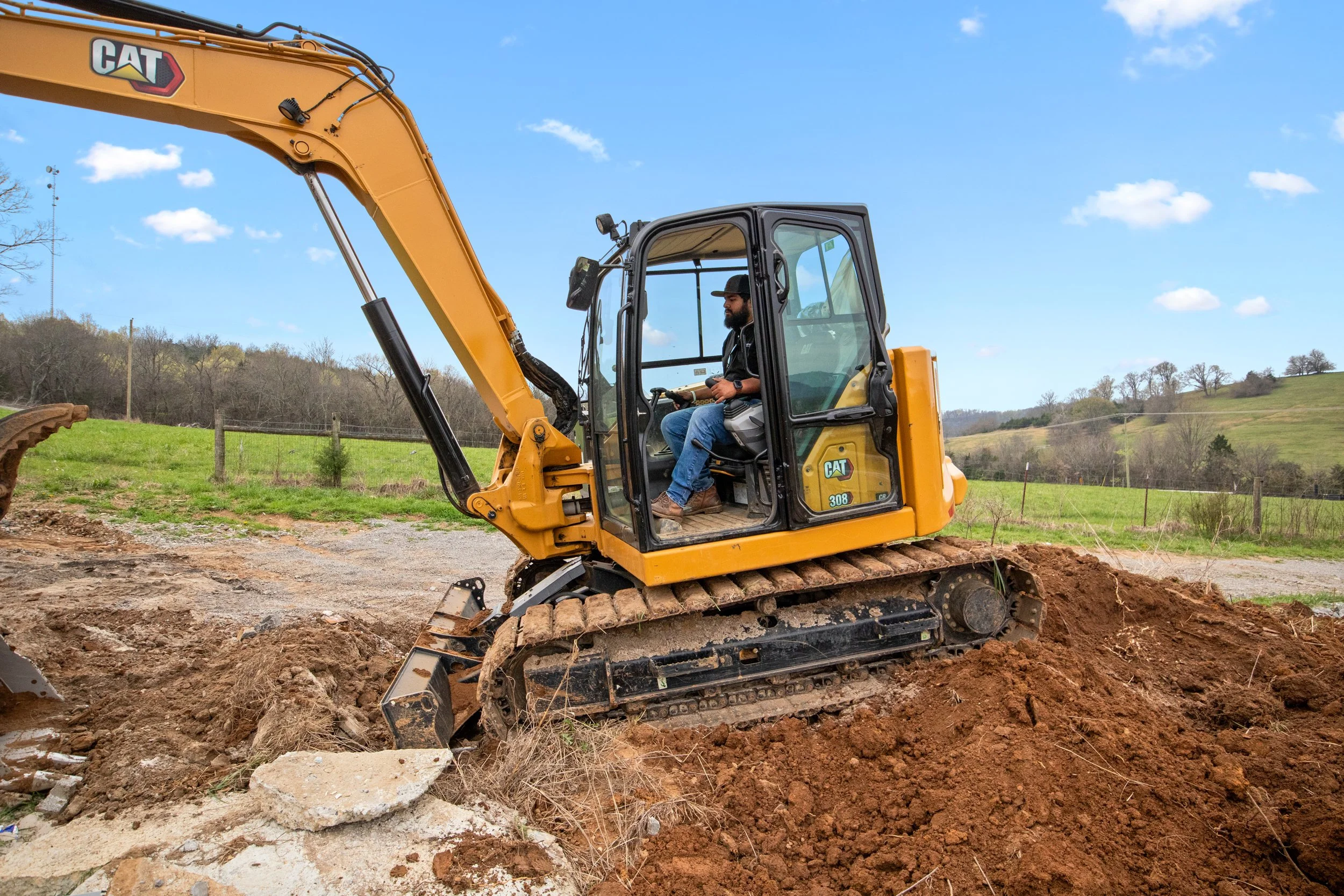 Francis Vega, of Vega Bros Construction, operating a yellow Caterpillar excavator at a construction site in a rural area with green fields and trees.