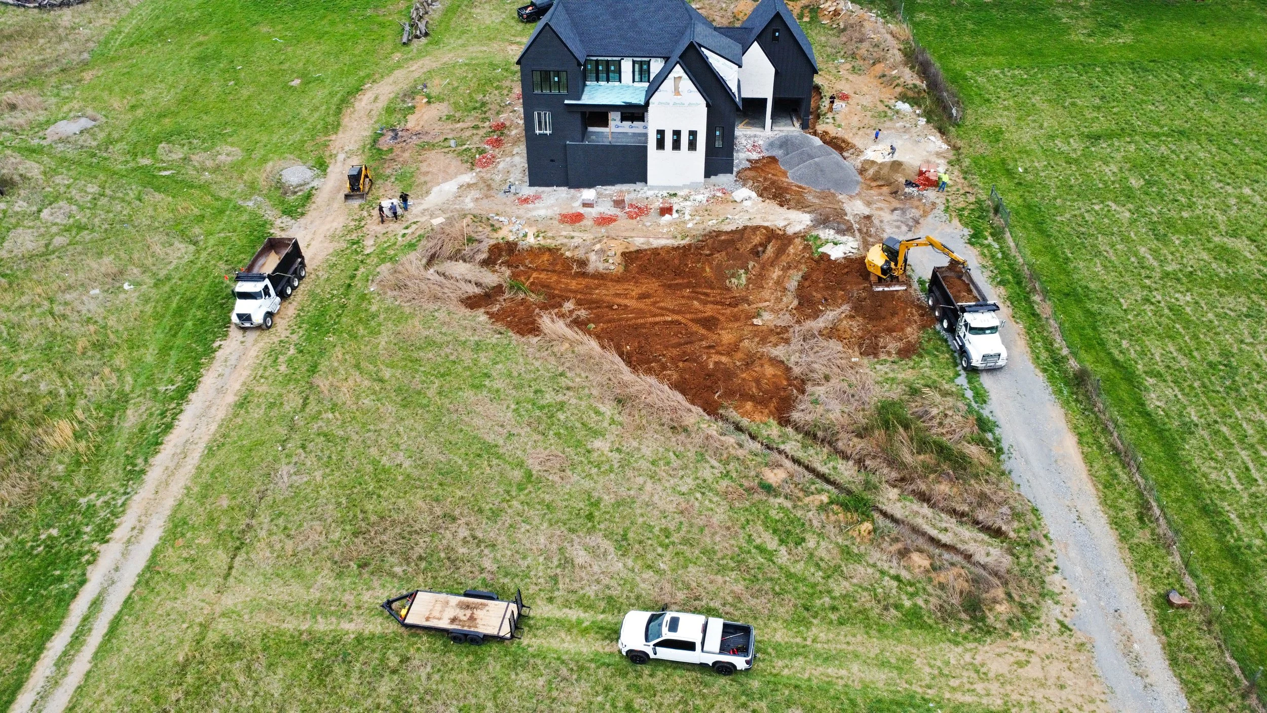 An aerial view of a house under construction with construction vehicles and workers present. There is a large excavation site in front of the house surrounded by green fields.