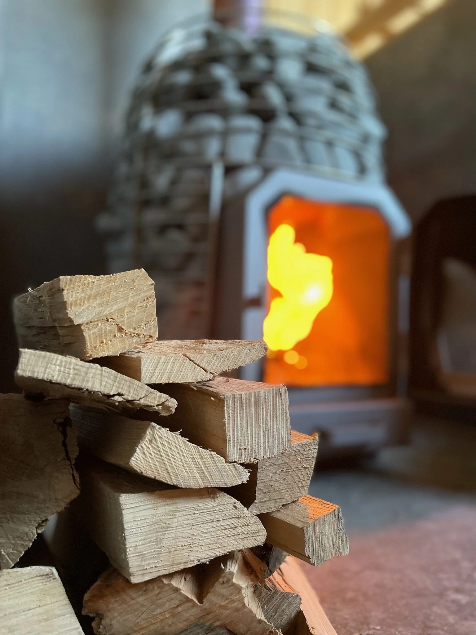Stack of chopped firewood in front of a lit wood stove with visible flames inside.