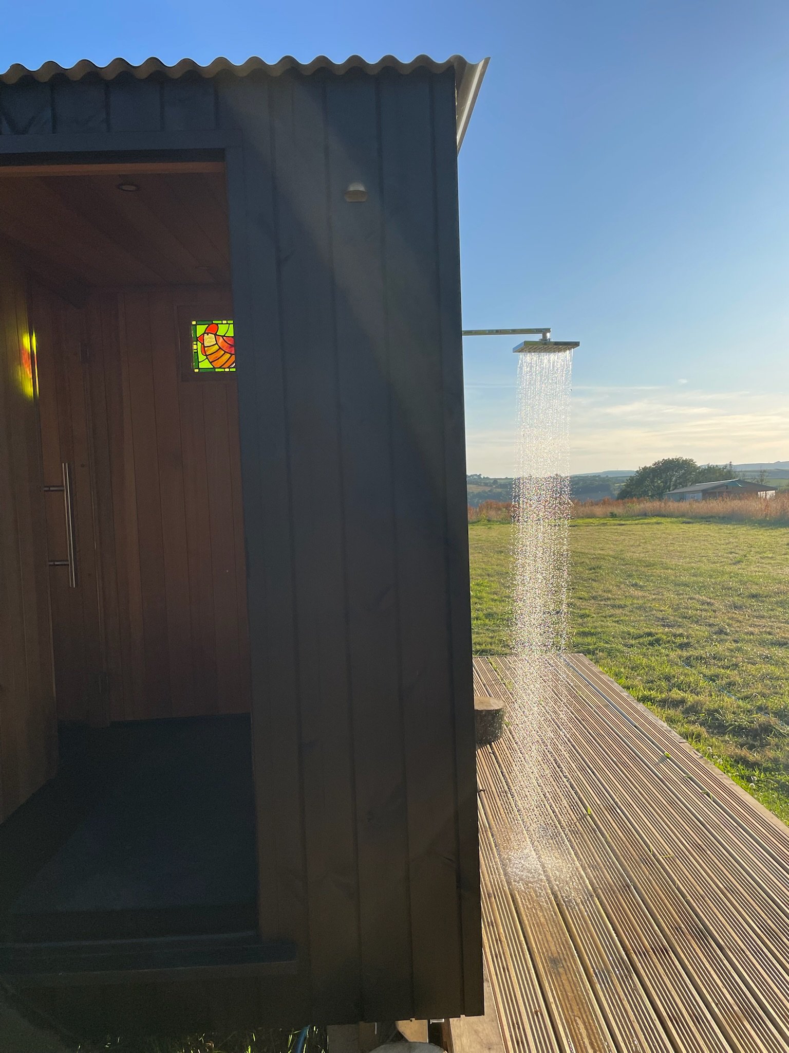 An outdoor shower with water flowing, attached to a dark wooden building with a stained glass window, on a wooden deck with a grassy field and blue sky in the background.