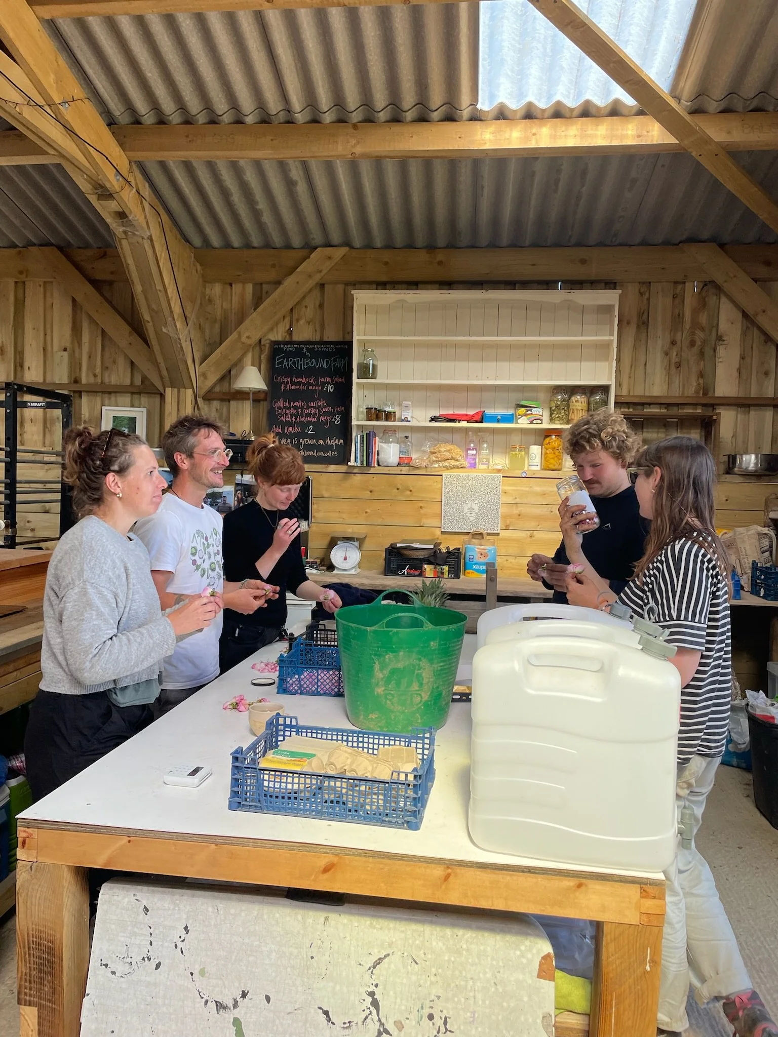 Group of five people gathered around a kitchen island in a rustic wooden barn, with a blackboard menu in the background.