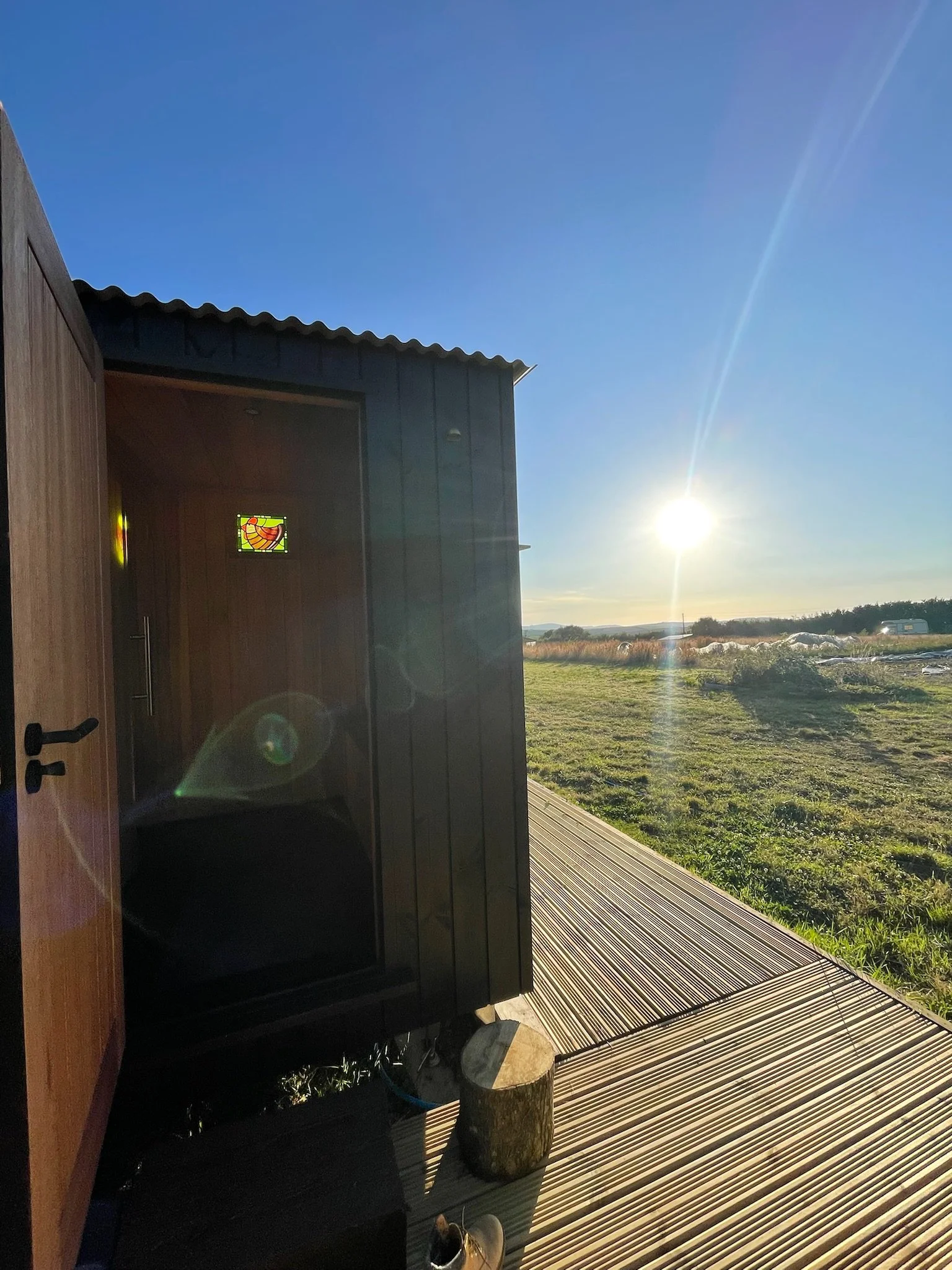 A small wooden building with an open door and a colorful stained glass window, next to a wooden deck on a sunny day with a clear blue sky and vast green fields in the background.