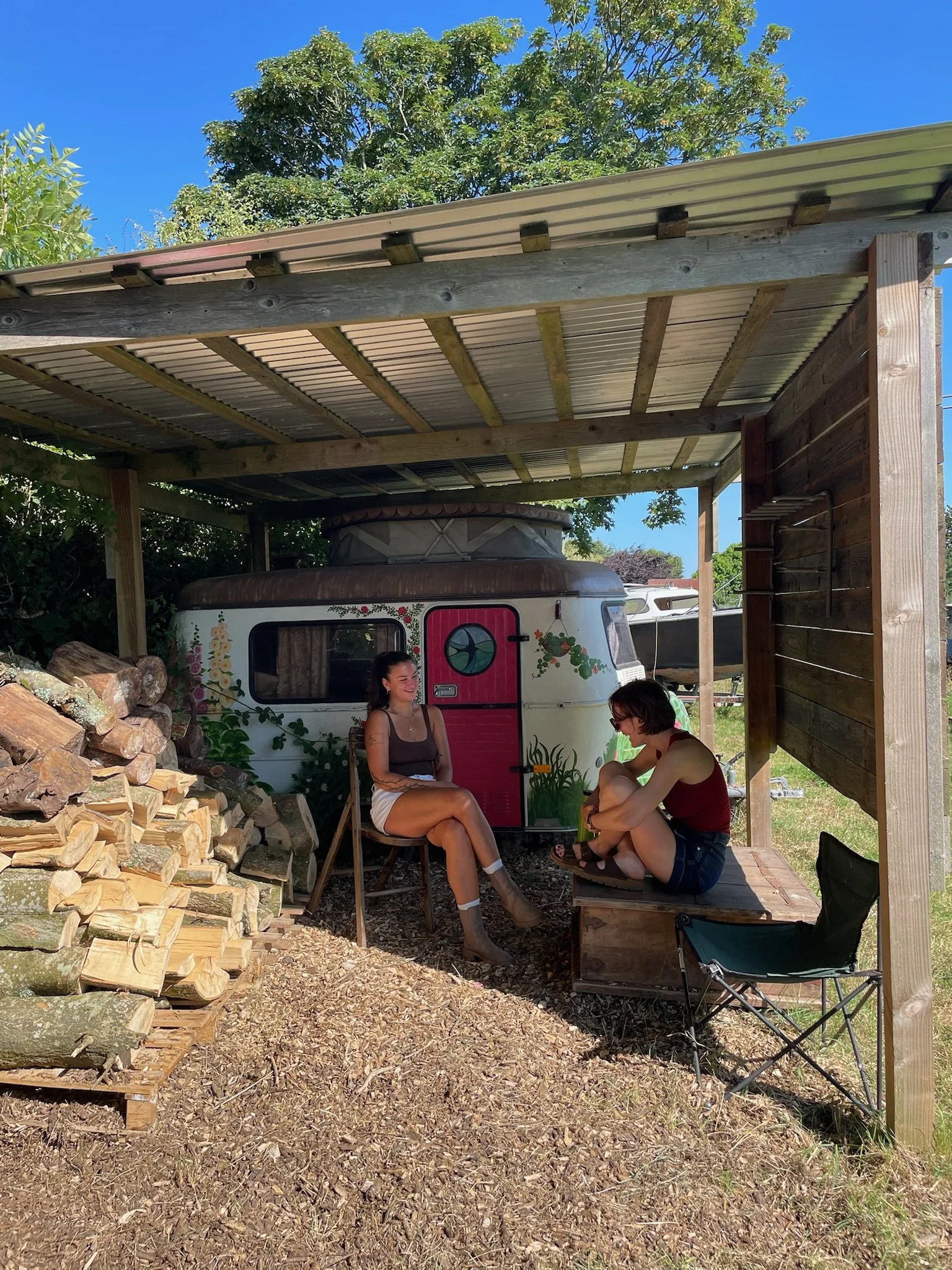 Two women sitting and chatting under a wooden shelter with a vintage camper decorated with flowers in the background, surrounded by wood logs.