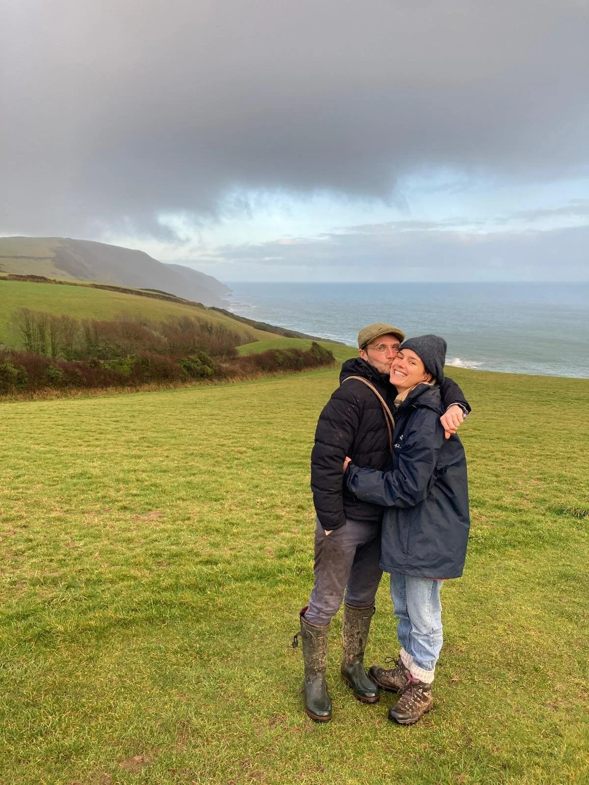 Two people hugging outdoors on a grassy field with green hills, coastline, and cloudy sky in the background.