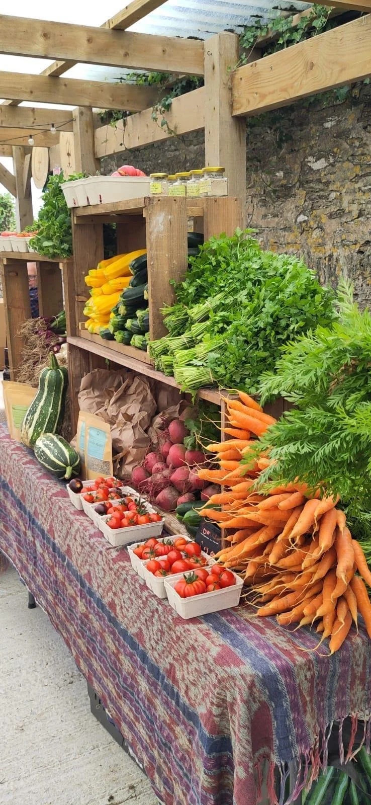 Fresh vegetables on display at a farmers market, including carrots, tomatoes, onions, zucchini, and squash, with a wooden structure and stone wall in the background.
