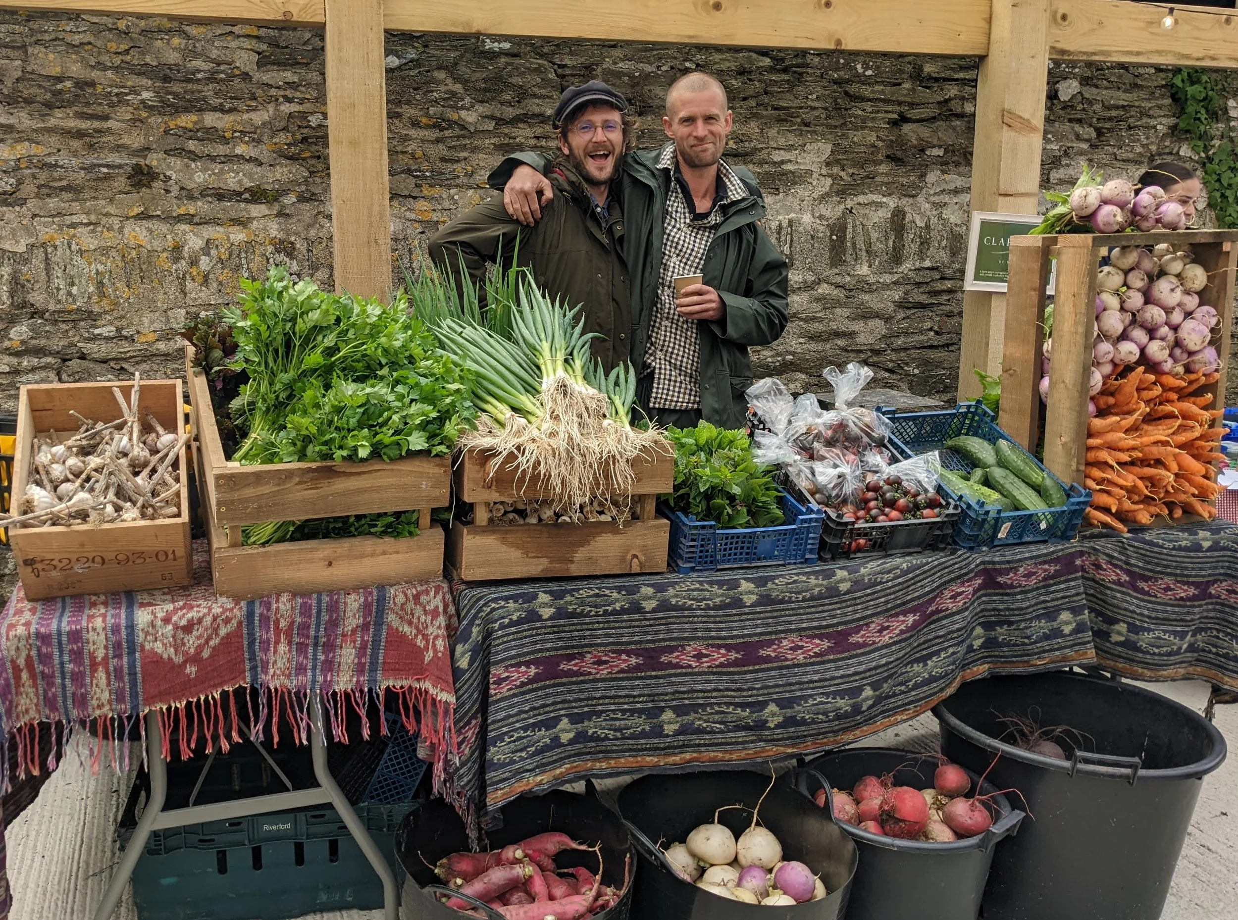 Two men standing behind a table filled with fresh vegetables at a farmers market. The table has garlic, greens, pineapples, tomatoes, cucumbers, and carrots. Buckets of radishes and turnips are in front. The men are smiling and one has his arm around the other, both dressed in jackets.