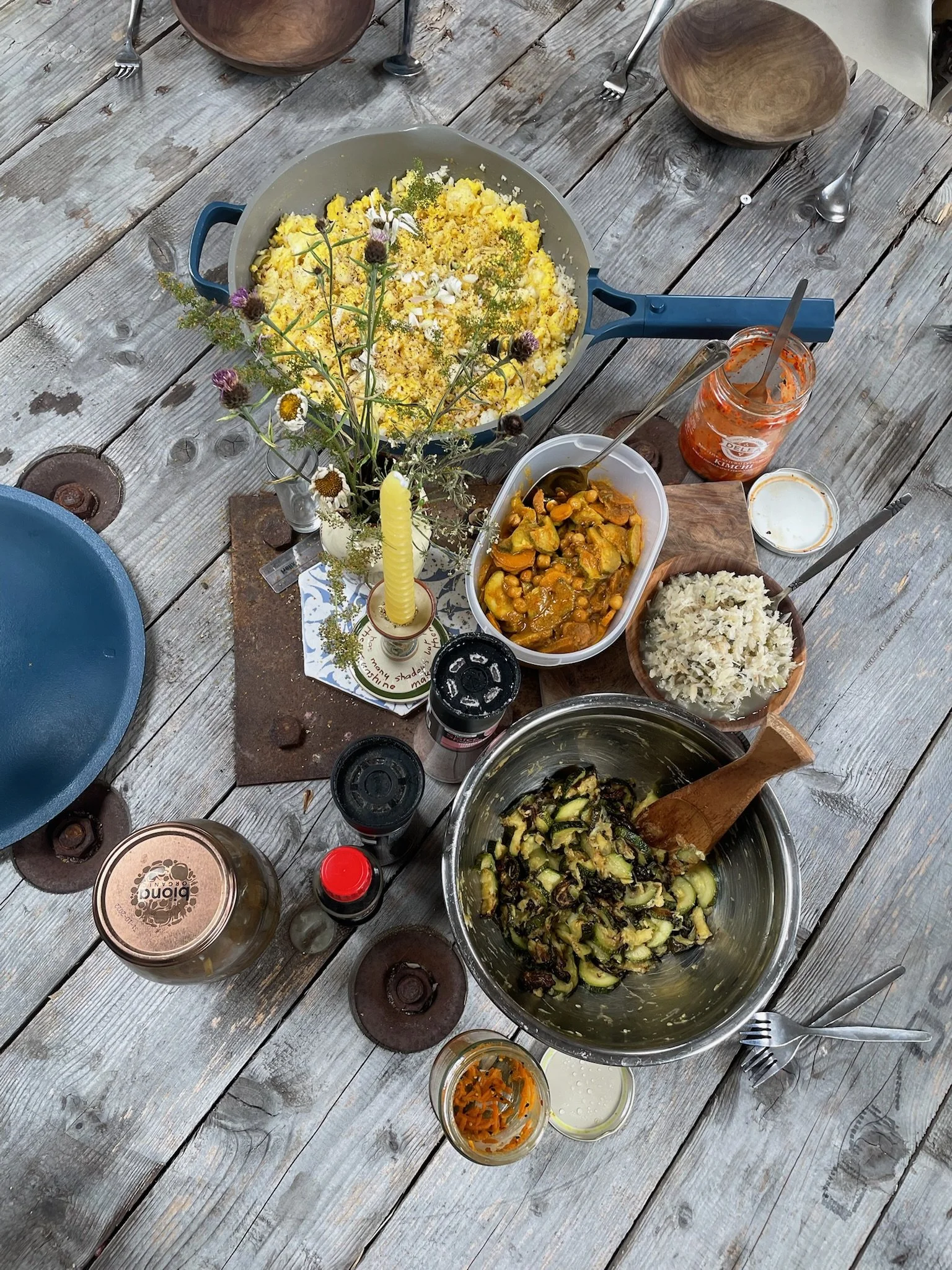 A supper club - rustic outdoor table set with various dishes, including scrambled eggs with herbs, a vegetable curry, rice, roasted zucchini, and condiments, along with a flower arrangement, a candlestick, and some spices.