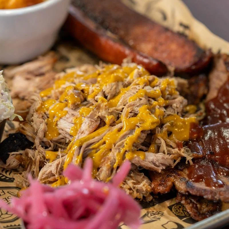 Close-up of a barbecue platter with shredded meat topped with mustard, grilled ribs, smoked sausage, and pulled pork with barbecue sauce, with pink pickled onions in the foreground.