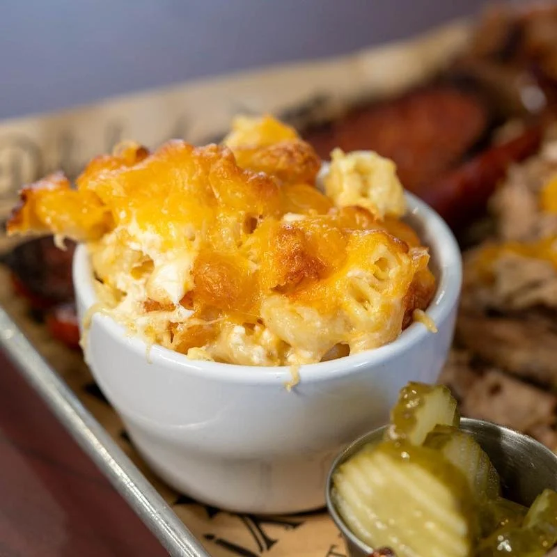 Baked macaroni and cheese in a white cup, with chopped pickles in a small metal container in the foreground, and bacon and grilled meat in the background.