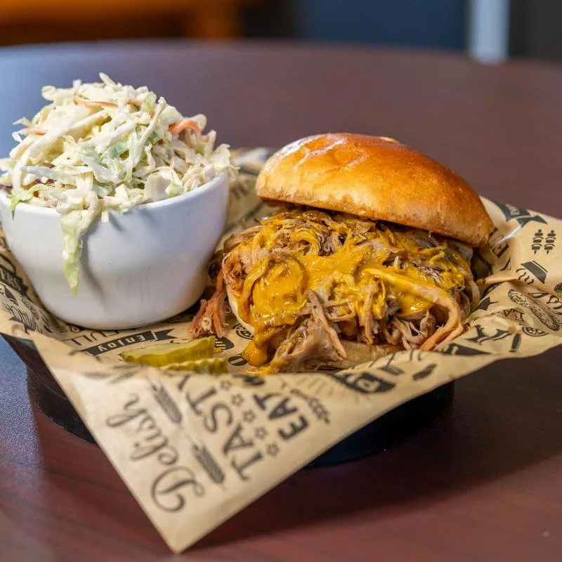Pulled pork sandwich with melted cheese on a toasted bun, served with coleslaw in a white bowl on a black tray with branded paper liner.