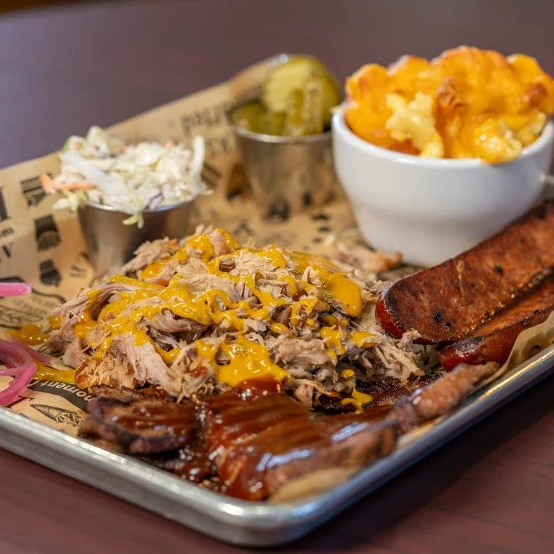 Barbecue platter with shredded pork topped with yellow mustard, baked macaroni and cheese, pickles, baked beans, ribs, and bacon-wrapped sausage on a tray.