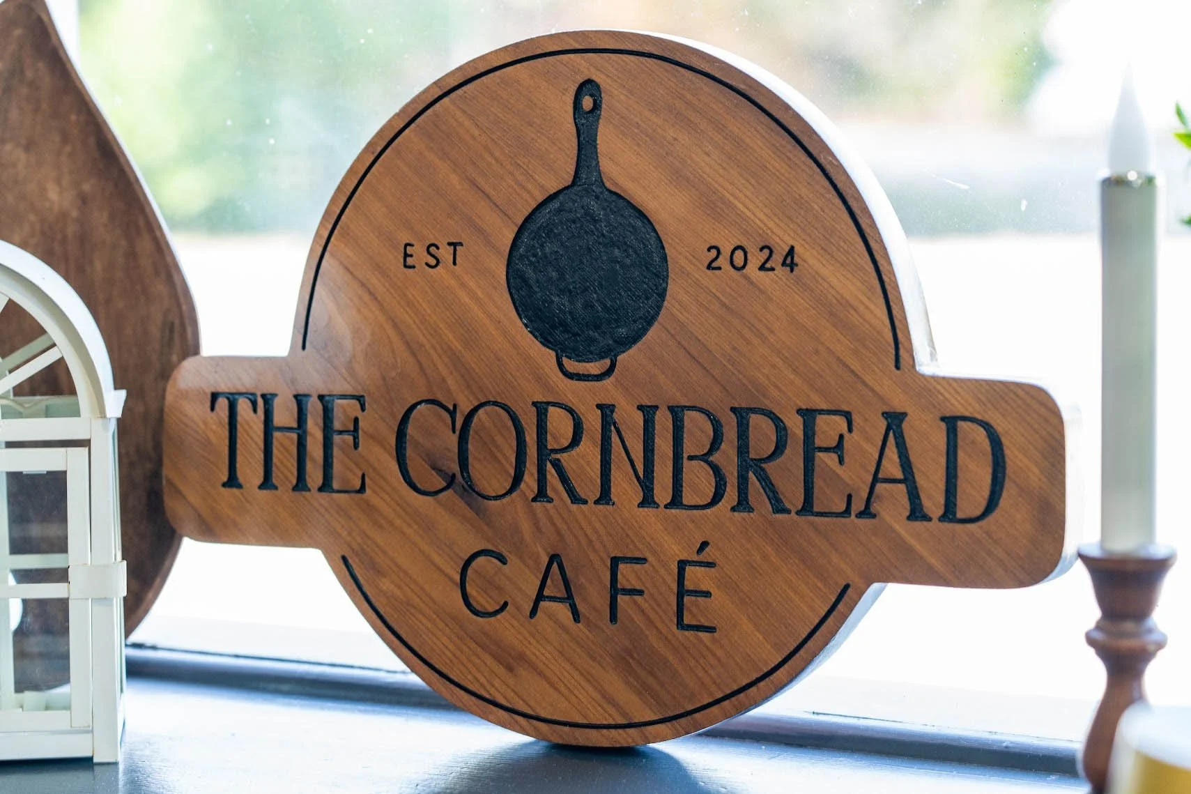 Wooden sign with a black skillet drawing, reading "The Cornbread Café," established in 2024, with decorative items on either side.