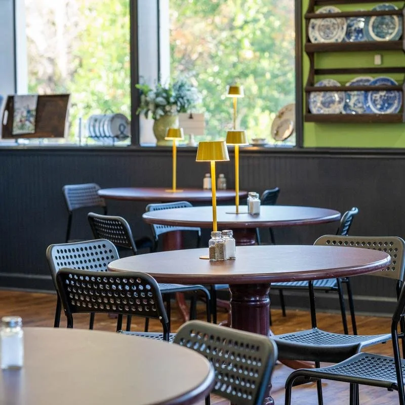 Empty dining area with round tables, black and tan chairs, yellow lamps, and a window view of greenery.