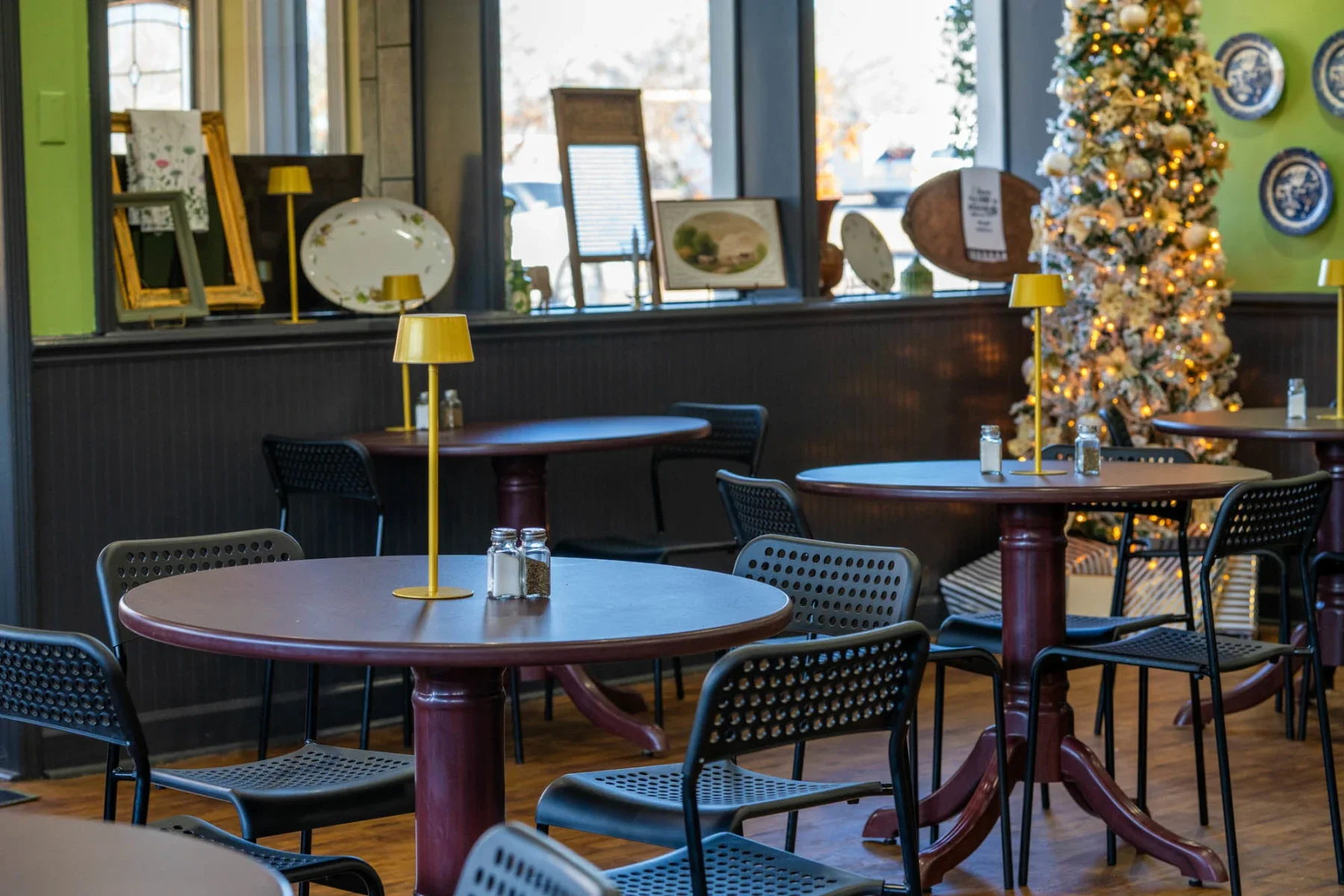 Restaurant with round wooden tables, black chairs, yellow lamps, salt and pepper shakers, and a decorated Christmas tree in the background.