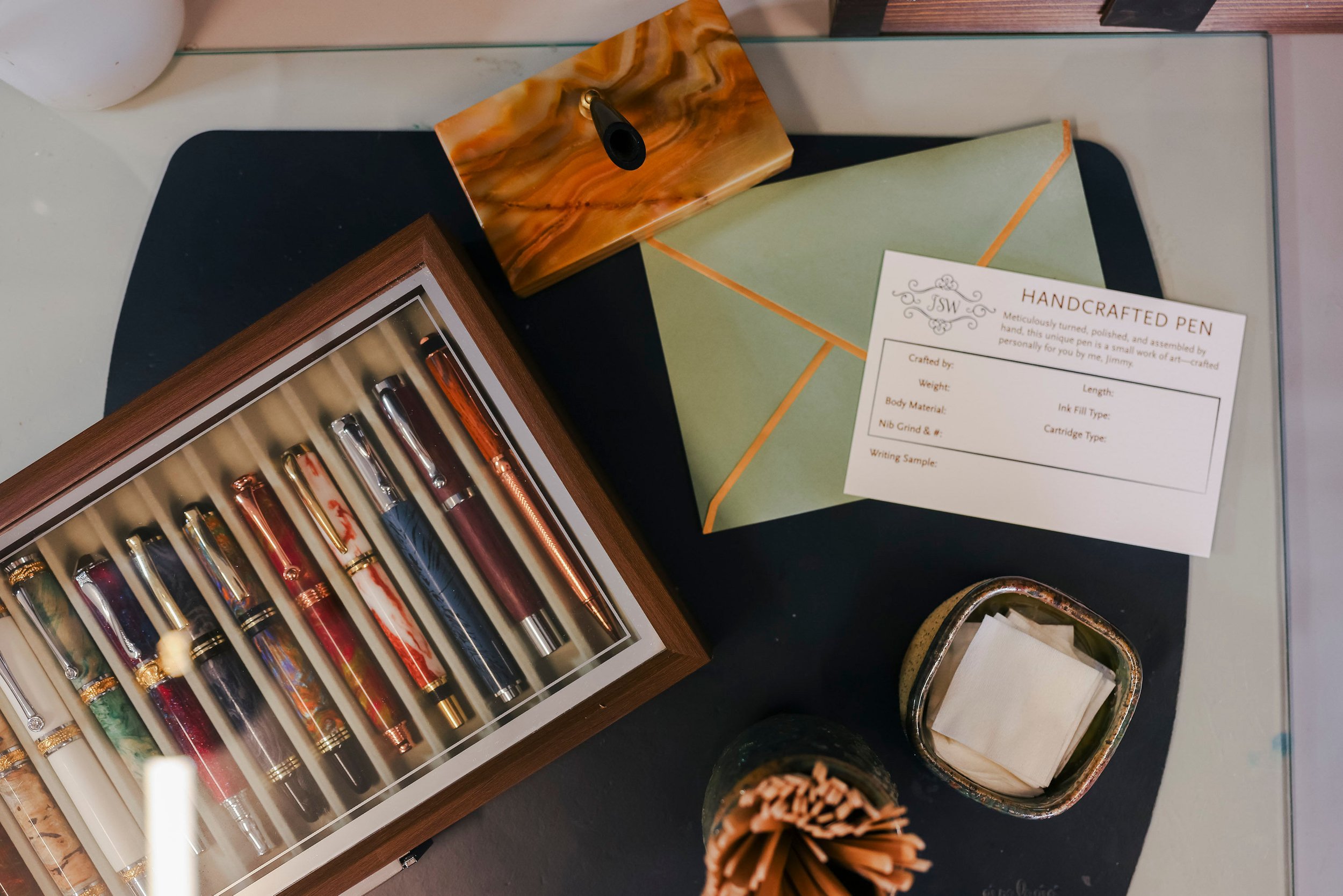 A display of various colorful fountain pens in a wooden box, a box with small sticks, a green and orange gift bag, and a small section of a black surface on which the items are placed.