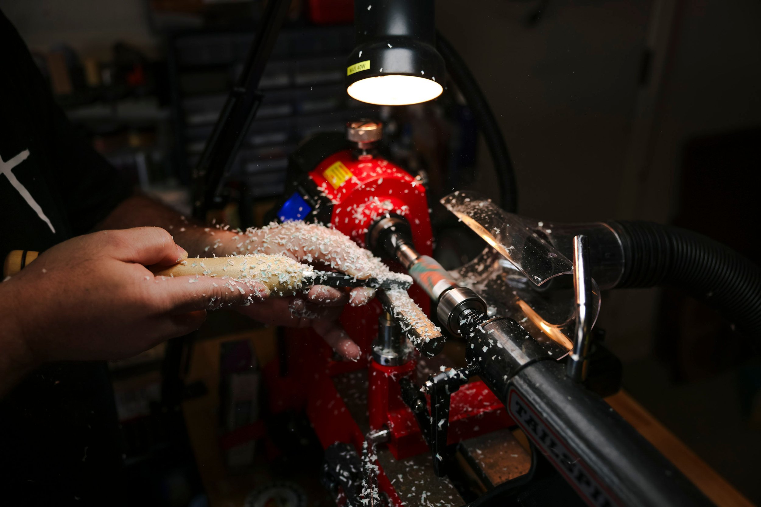 A person operating a wood lathe machine, shaping a piece of wood with wood shavings scattered around, in a workshop setting.