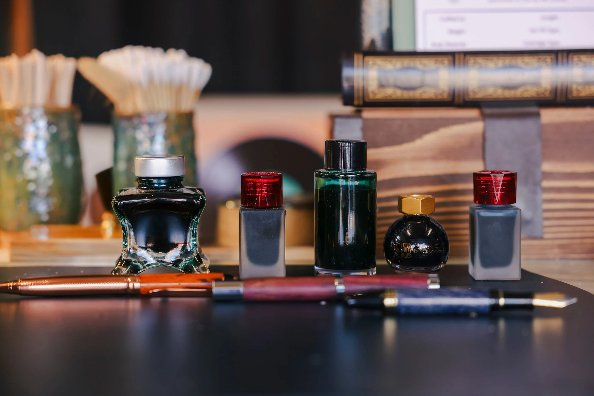 Close-up of various perfume bottles on a wooden surface with blurred jars and a computer screen in the background.