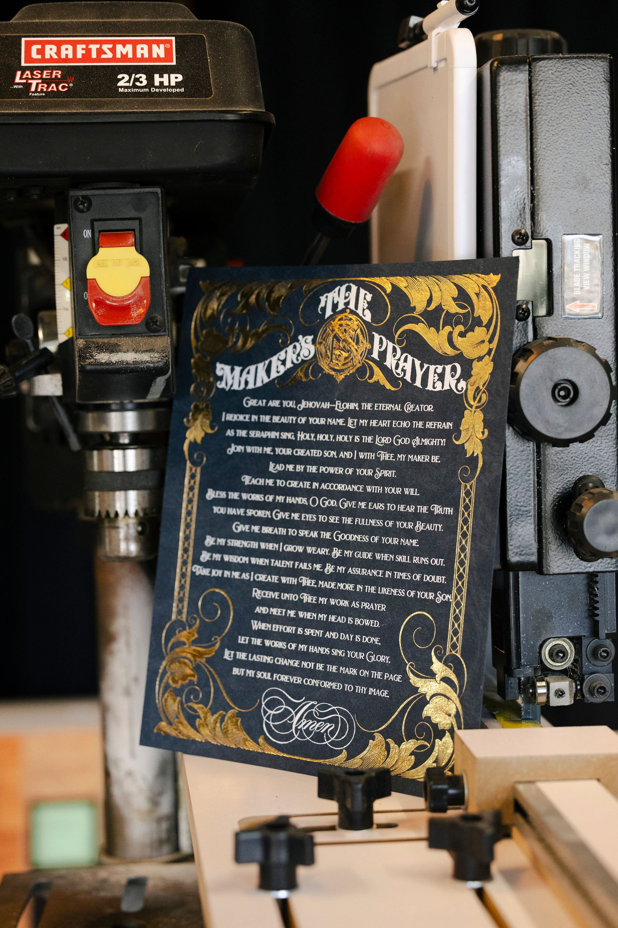 A woodworking saw machine with a museum-style prayer card attached to the machine. The prayer card has gold and white lettering with decorative gold and black borders, featuring a prayer starting with 'The Makers Prayer.'