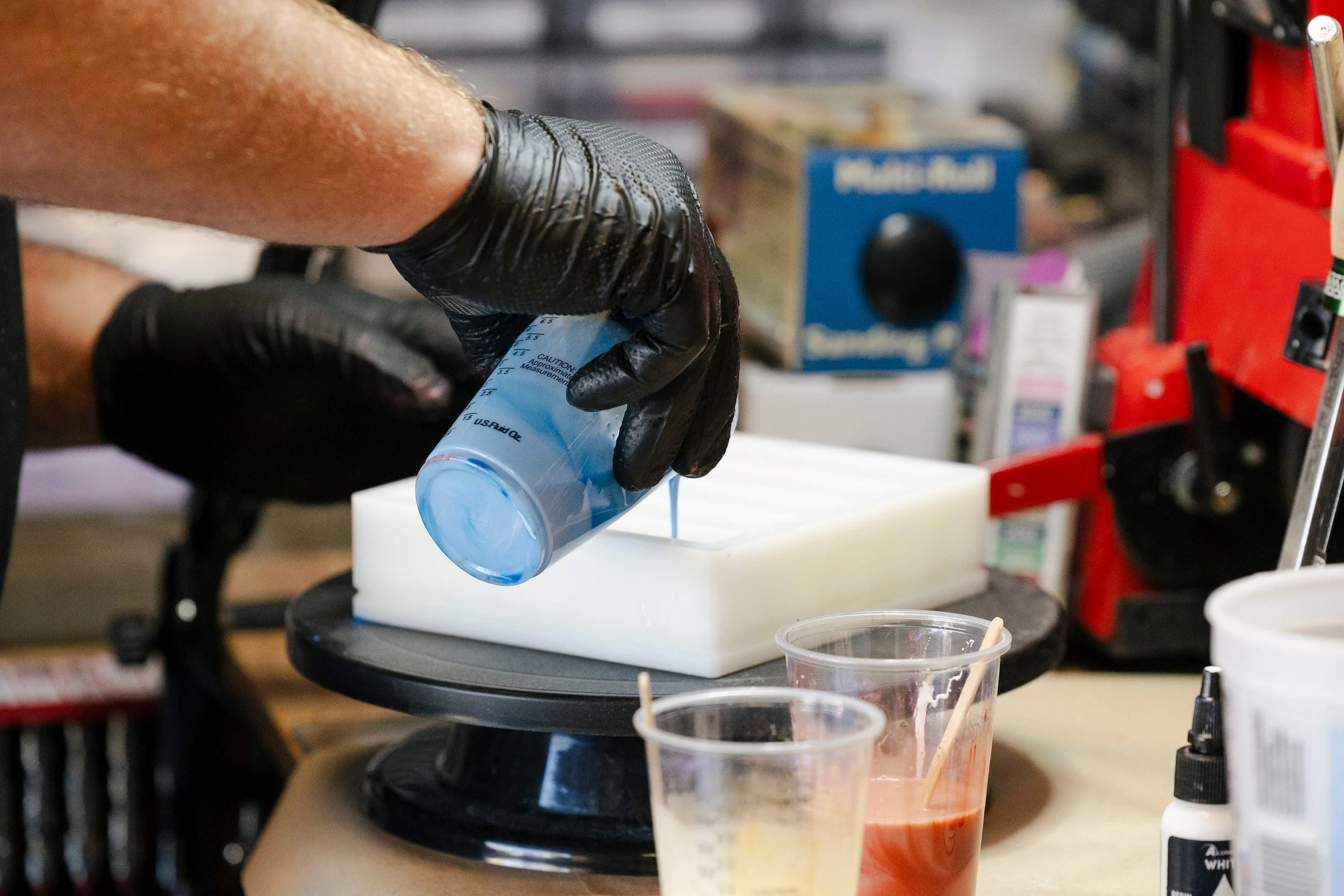 Person wearing black gloves pouring a blue liquid into a white mold in a workshop or laboratory setting.