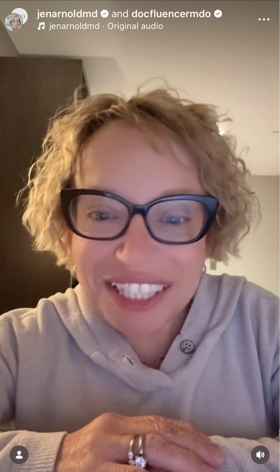 A woman with short curly blond hair and wearing glasses is smiling and looking down. She is wearing a light gray hooded sweatshirt and has a ring on her left hand, resting on a table or surface.